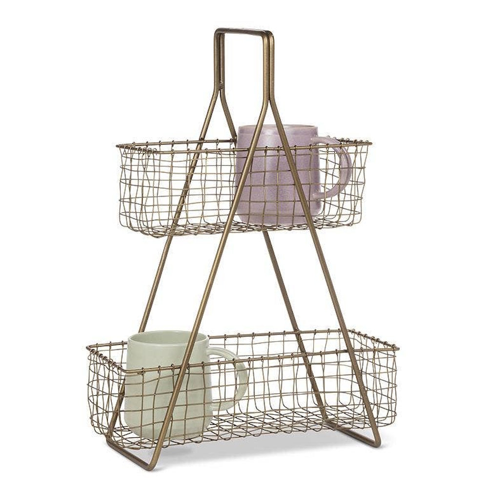 Two-tiered brass wire organizer stand with top and bottom baskets, each holding a ceramic mug.