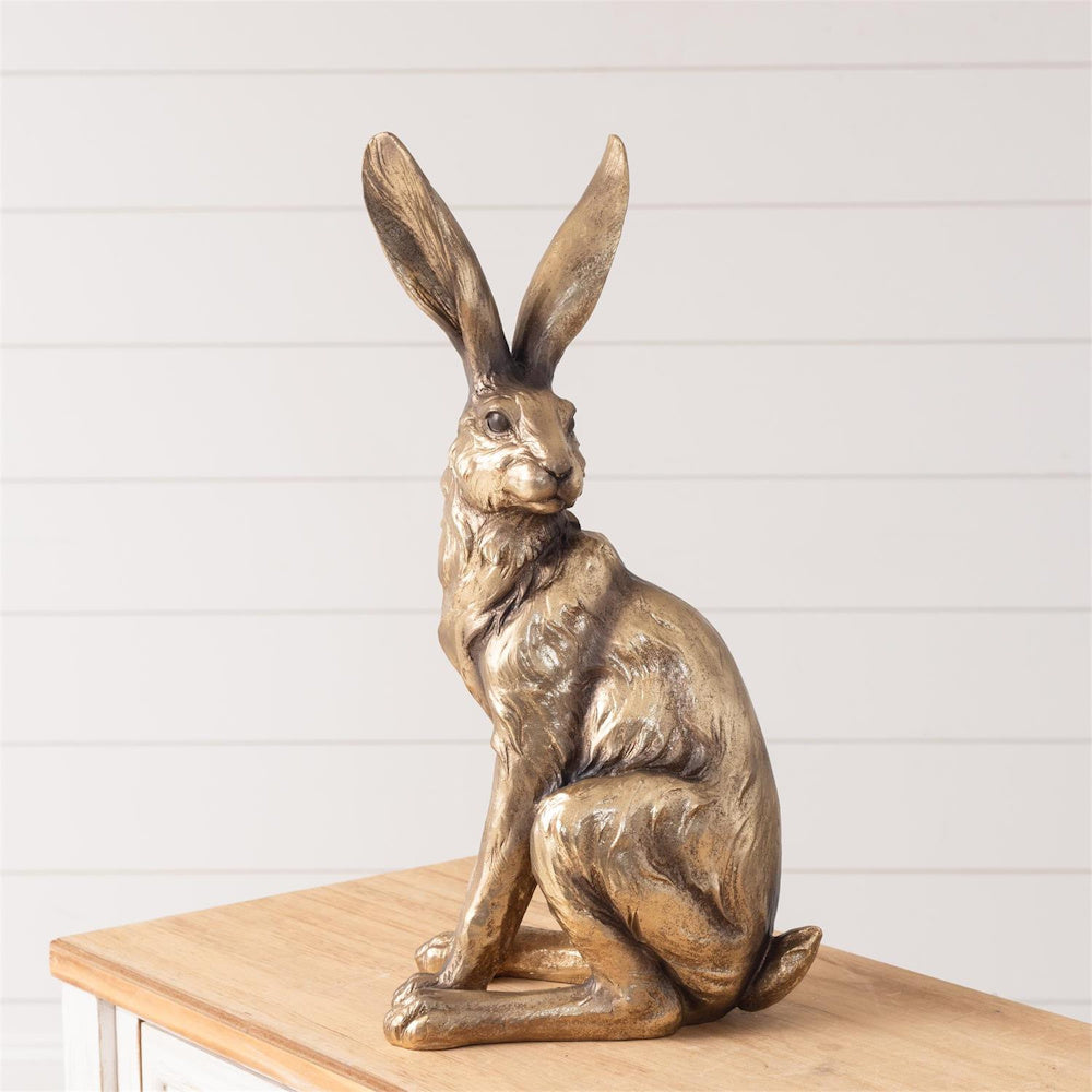 Antique gold standing hare statue with long ears, sitting on a wooden surface against a white shiplap wall.