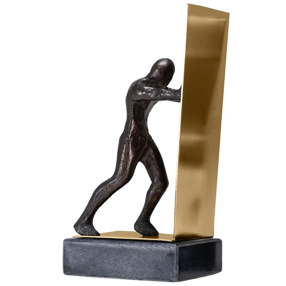 Dark metallic sculpture of a man pushing a vertical gold metal sheet, set on a dark rectangular base.
