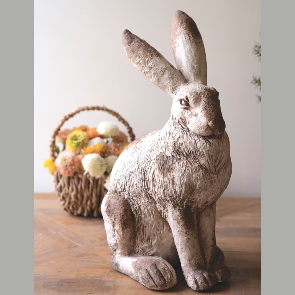 Rustic faux concrete rabbit statue sitting on a wooden table next to a woven basket of colorful spring flowers.