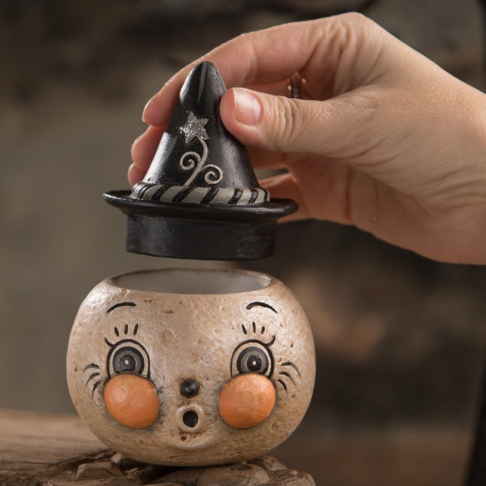 A hand lifts a black witch hat lid off a round, vintage-style Halloween container with a surprised face.