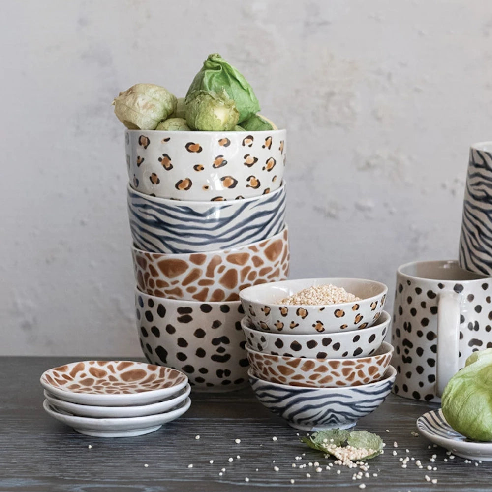 Stacked ceramic bowls and mugs with animal prints like leopard, zebra, and giraffe on a rustic wooden table.