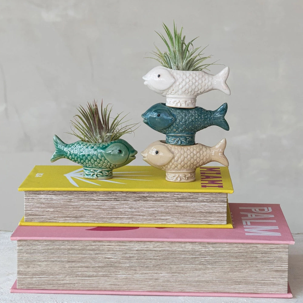 Stacked ceramic fish air plant holders in white, blue, and tan on colorful books.