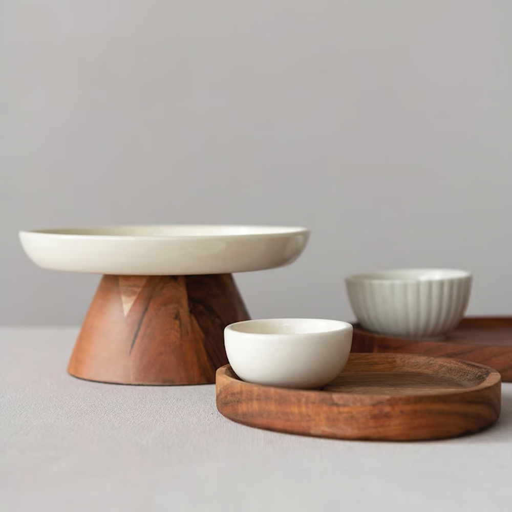 Cream stoneware footed pedestal with a natural acacia wood base, displayed alongside wooden trays and ceramic bowls.