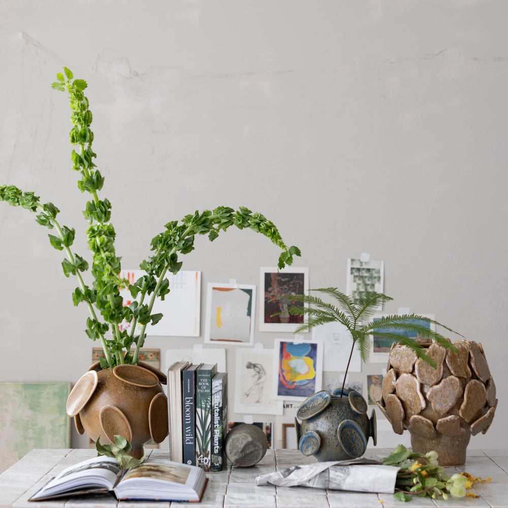 Three unique stoneware planters with 3D designs displaying greenery on a table with books and artwork.