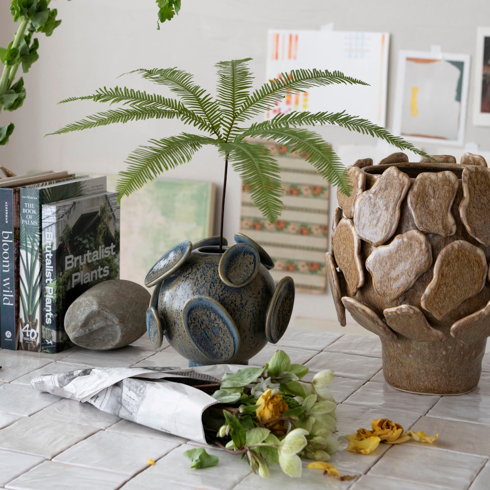 A blue textured vase holding a fern frond sits beside a brown petal-like planter on a tiled tabletop.
