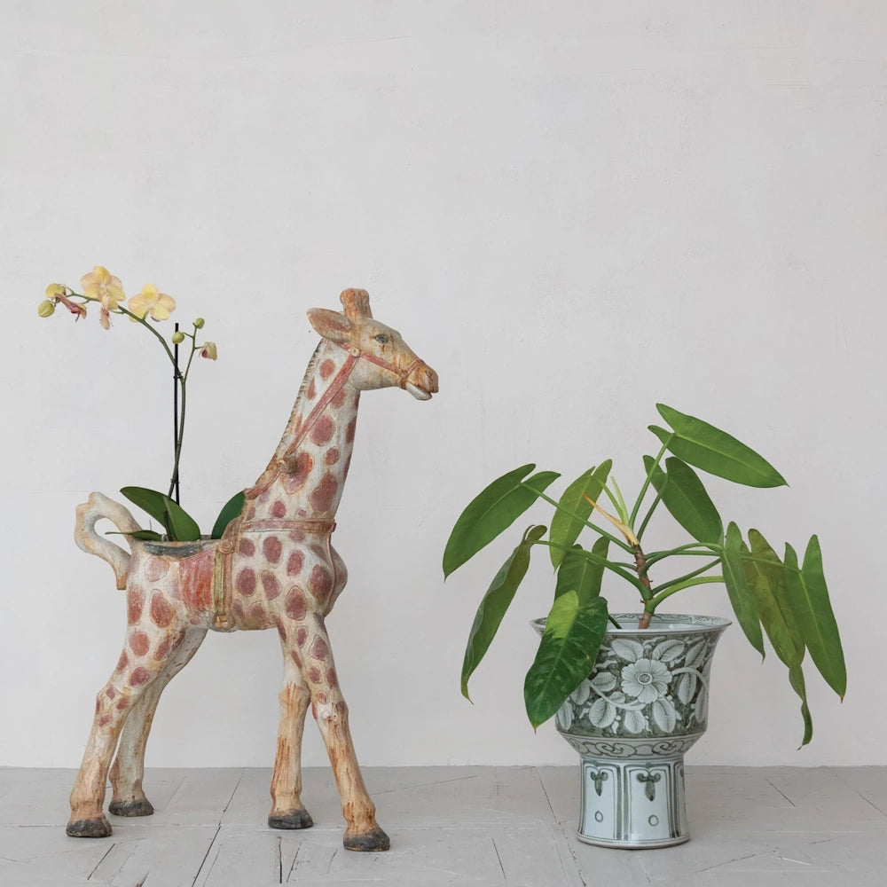 A hand-painted giraffe planter with yellow orchids next to a stoneware floral footed planter holding a green leafy plant.