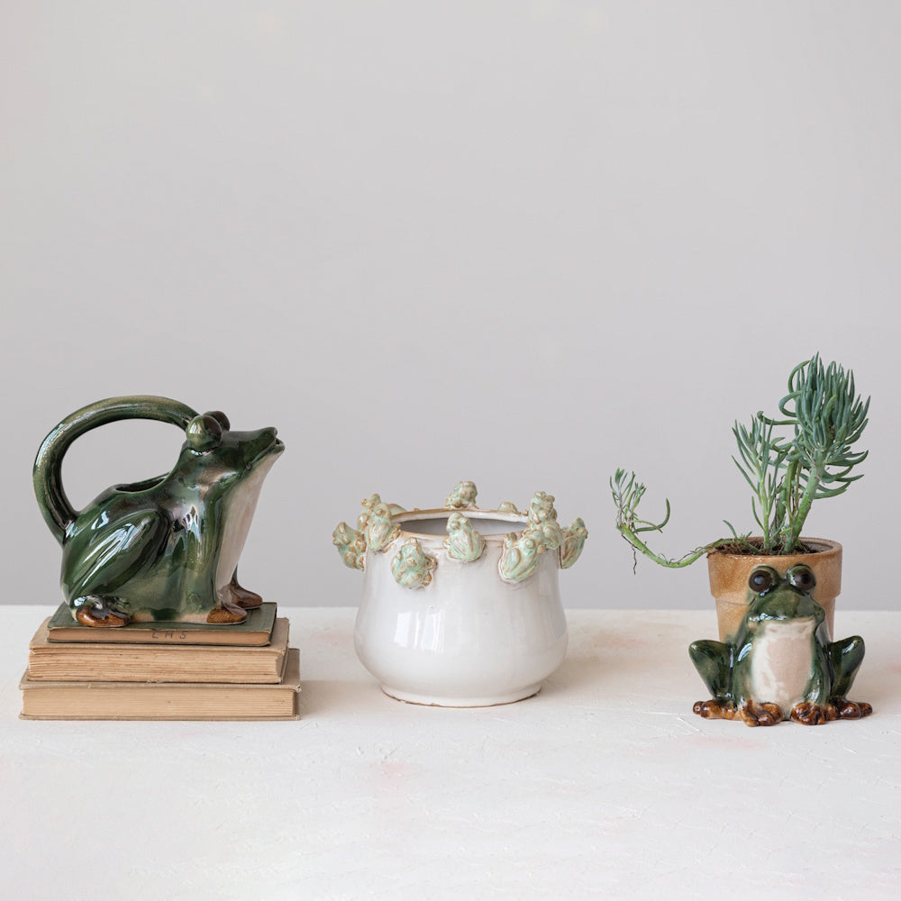 Three frog-themed ceramic items, including a pitcher, a planter, and a small figure next to a potted plant.