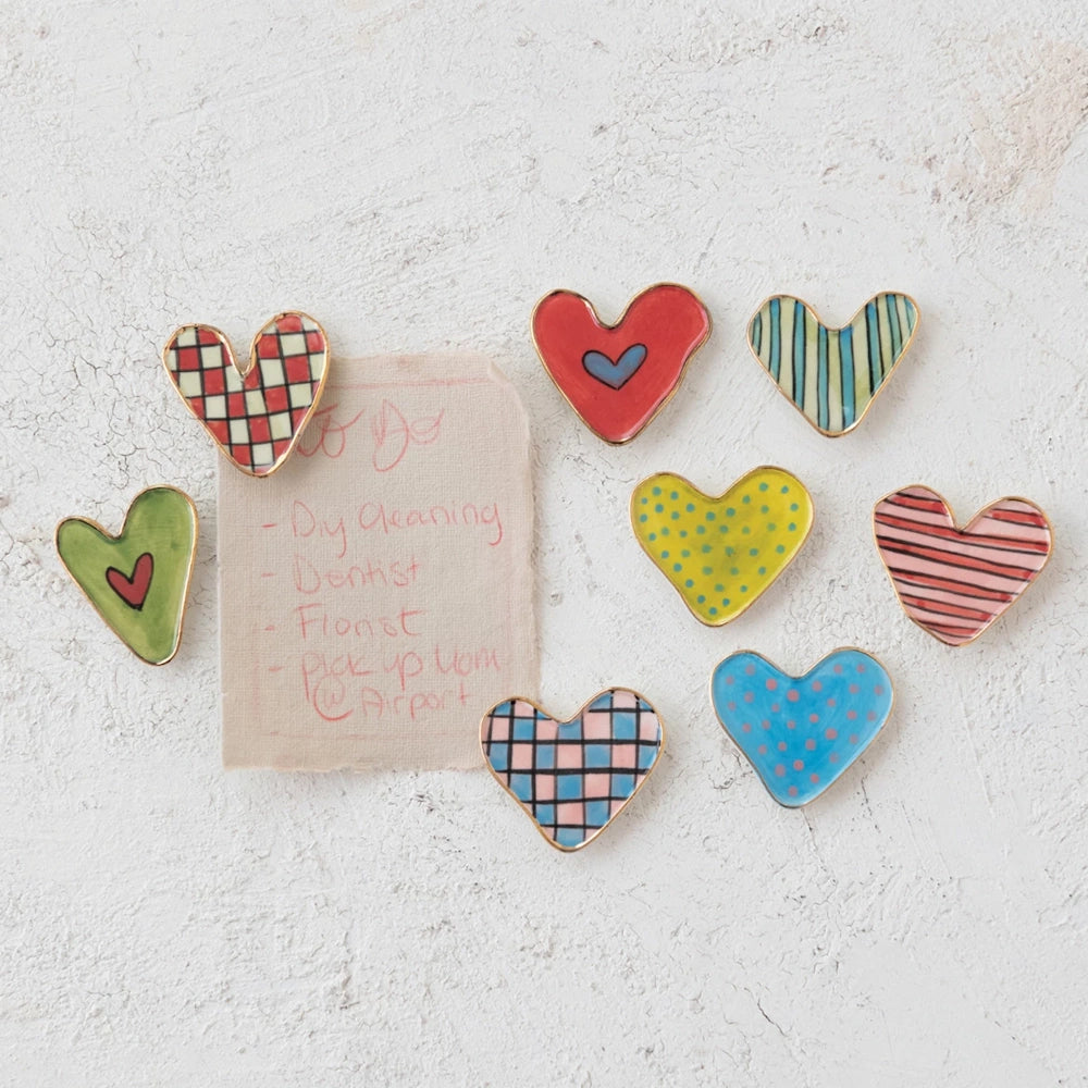 Eight colorful, patterned stoneware heart magnets with gold rims arranged on a white textured wall around a small note.