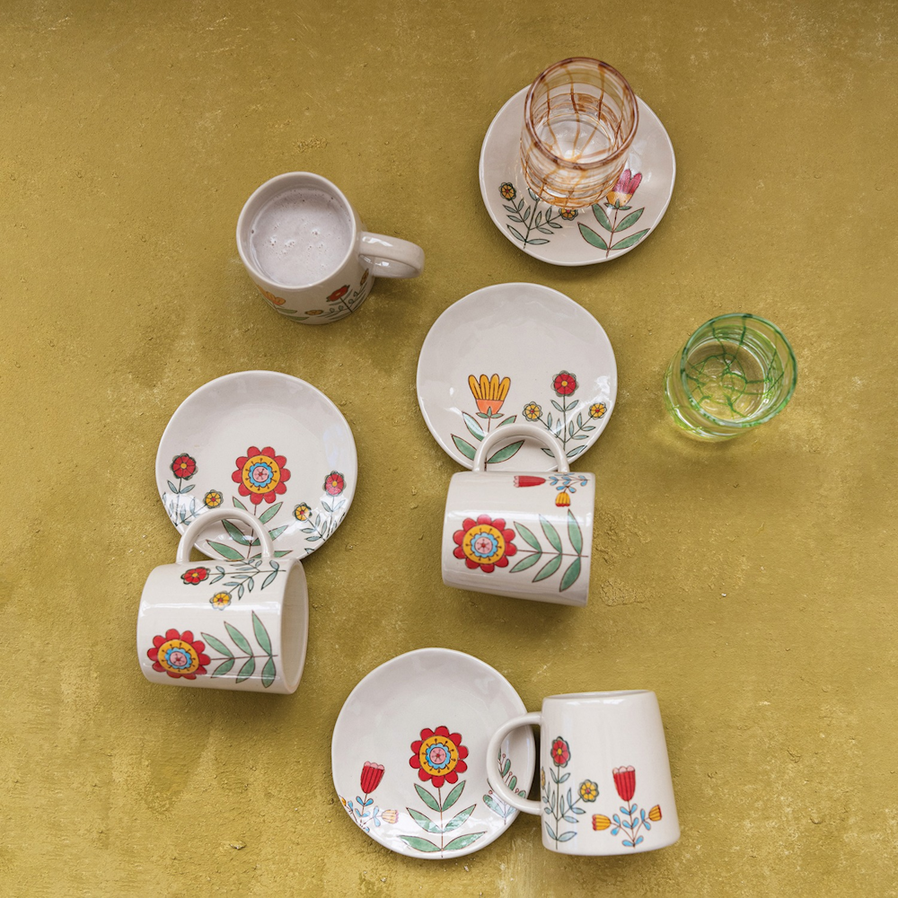 Hand-painted stoneware mugs and saucers with colorful floral patterns on a textured yellow backdrop.