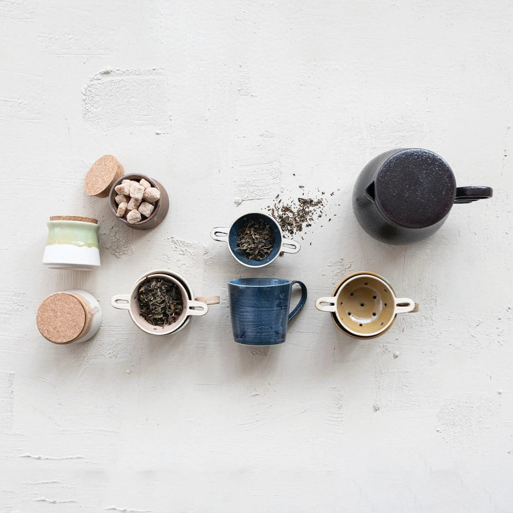 Flat lay of handmade stoneware tea set with blue mug, ceramic strainers, loose leaf tea, sugar cubes, and teapot.