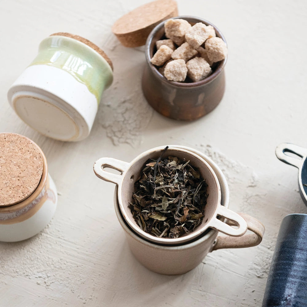 Stoneware tea mug with strainer, loose leaf tea, cork-lid jars, and brown sugar cubes on a neutral surface.