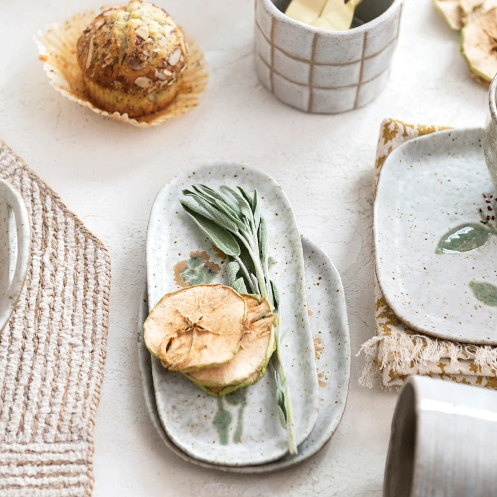 Speckled ceramic tray holding sage leaves and dried apple slices, set on a styled tabletop with pastries.