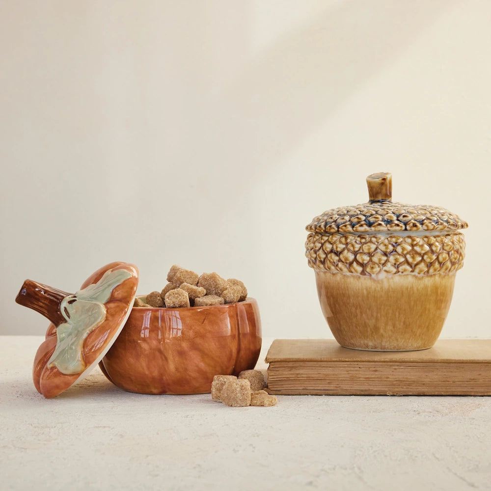 Stoneware pumpkin jar filled with brown sugar cubes next to a ceramic acorn jar on an old book.