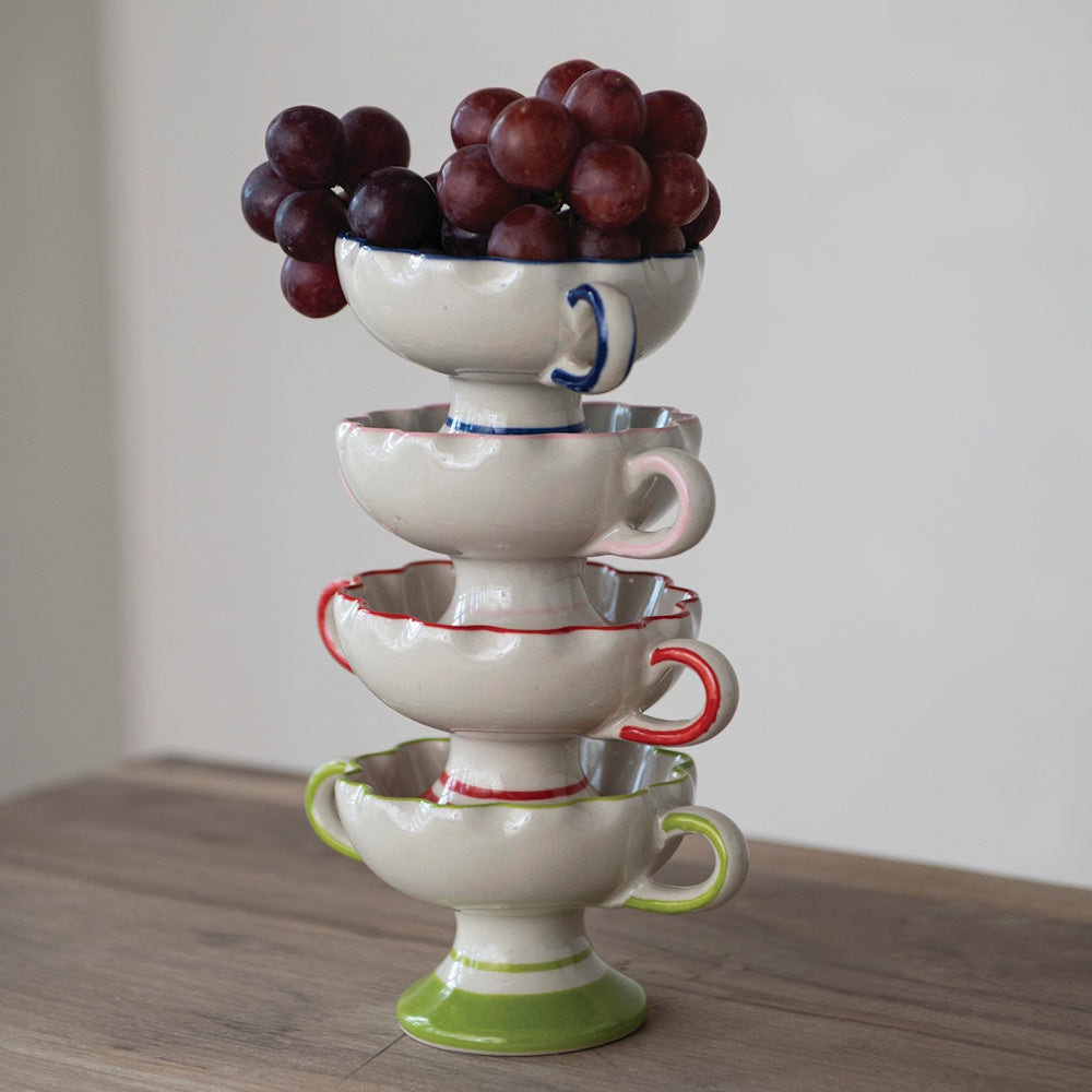 Four colorful scalloped footed bowls stacked vertically on a wooden table, topped with a bunch of red grapes.