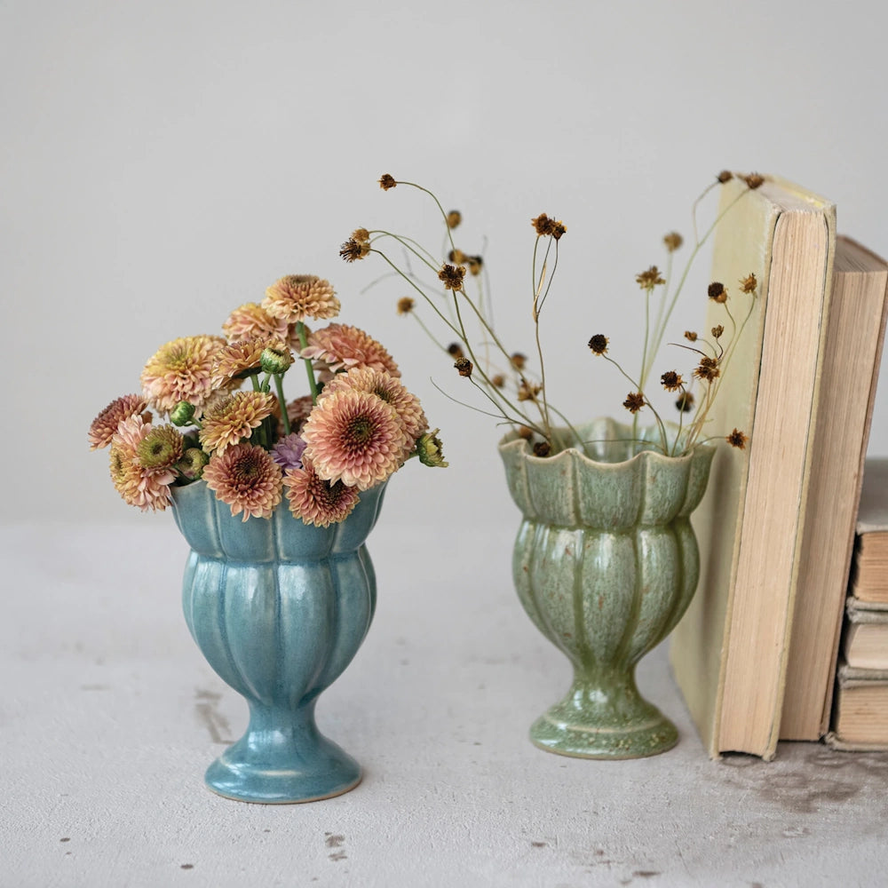 Blue ceramic footed vase with pink flowers next to a green vase with dried stems and old books.