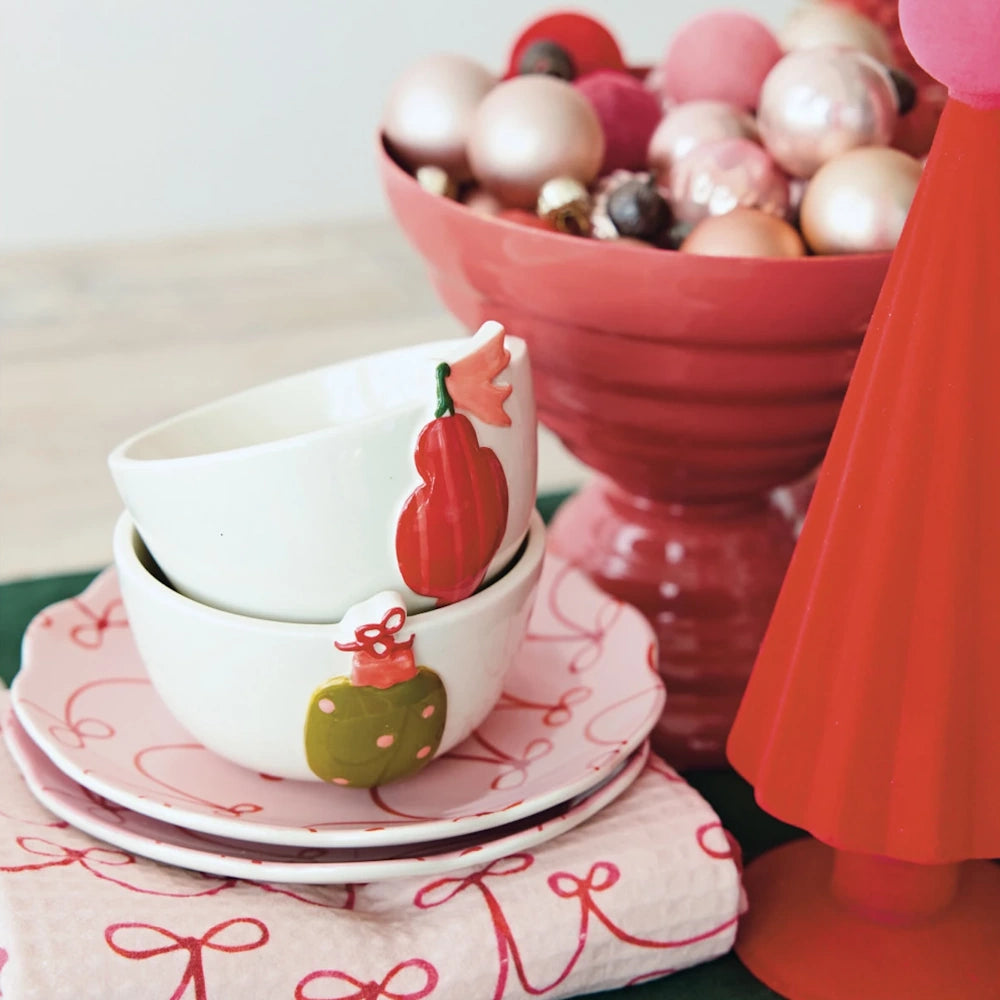 Two white ceramic ornament bowls stacked on pink bow-patterned plates next to festive holiday decor.
