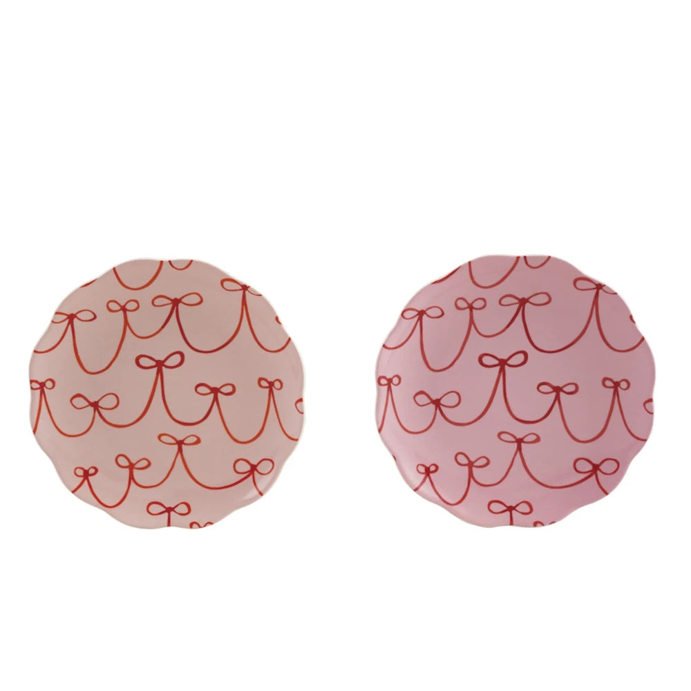 Two pink round scalloped ceramic plates with a red bow pattern side by side on a plain white background.