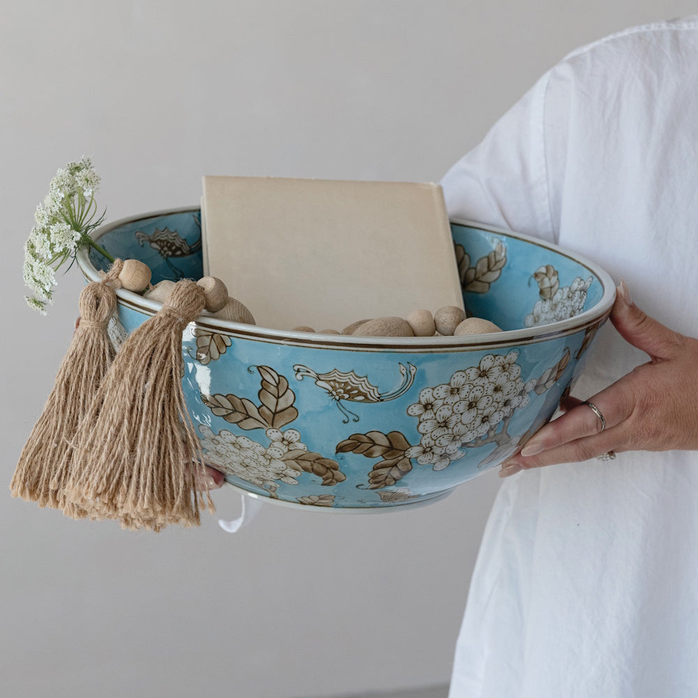 Blue stoneware bowl with white cherry blossoms and brown leaves, held by a person and styled with a book and tassels.