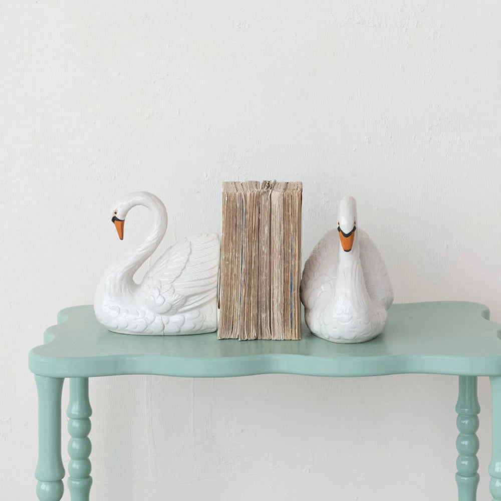 White ceramic swan bookends holding vintage books on a teal scalloped console table against a white wall.