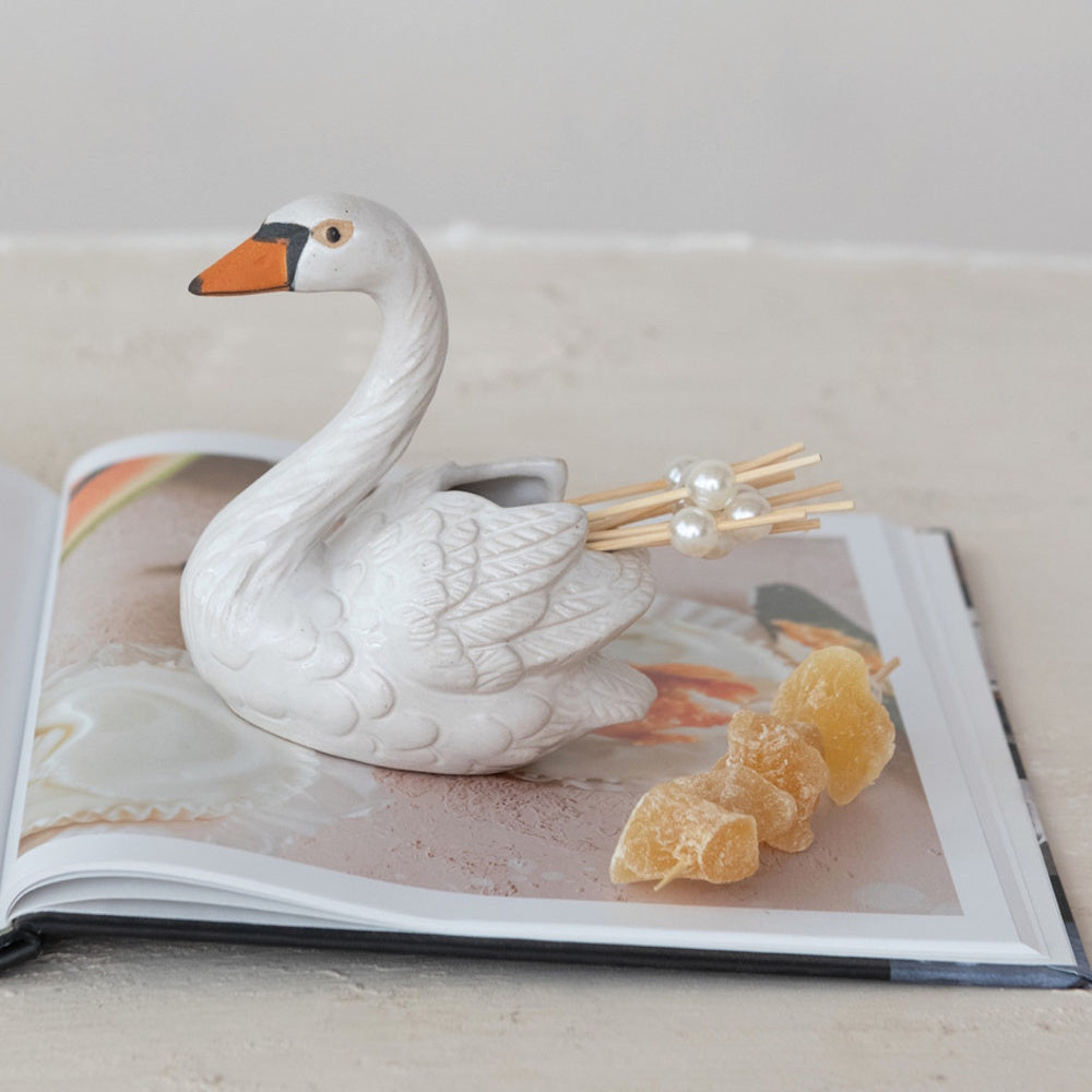 White stoneware swan-shaped toothpick holder with pearl-topped toothpicks, resting on an open book.