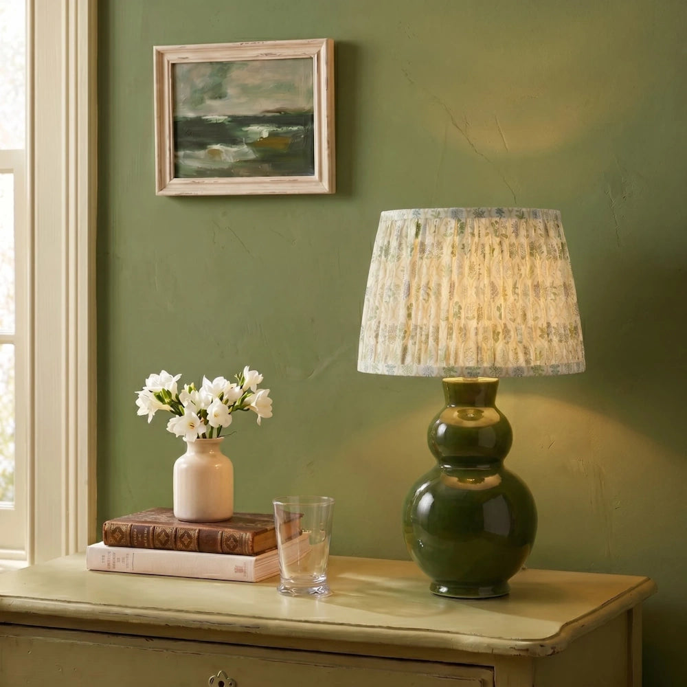 Lifestyle view of a dark green stoneware table lamp with a pleated floral fabric shade on a wooden cabinet next to flowers.