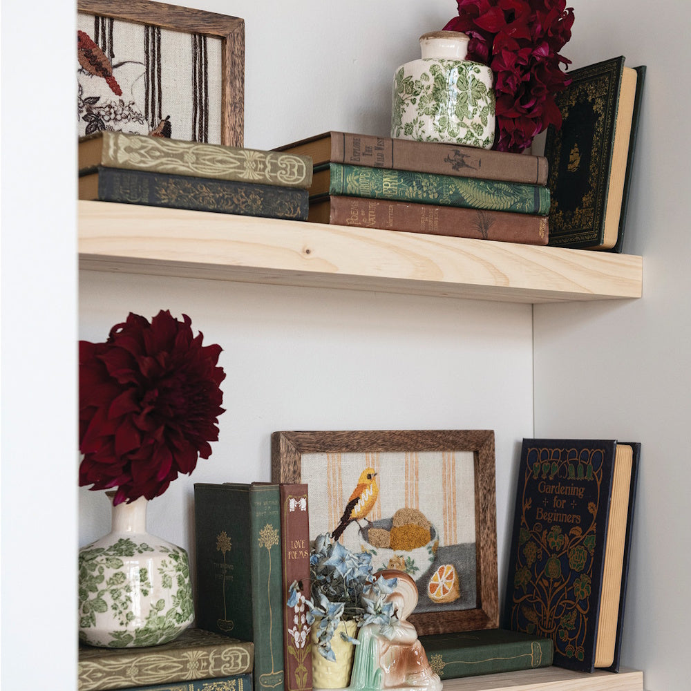 Styled wooden shelves with vintage books, framed art, floral patterned vases, and dark red flowers.