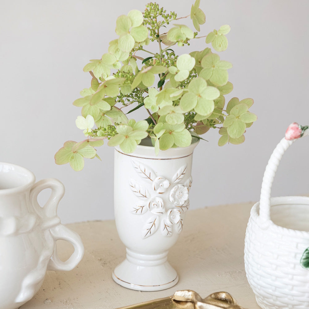 White vintage reproduction footed vase with raised floral details holding green hydrangeas.
