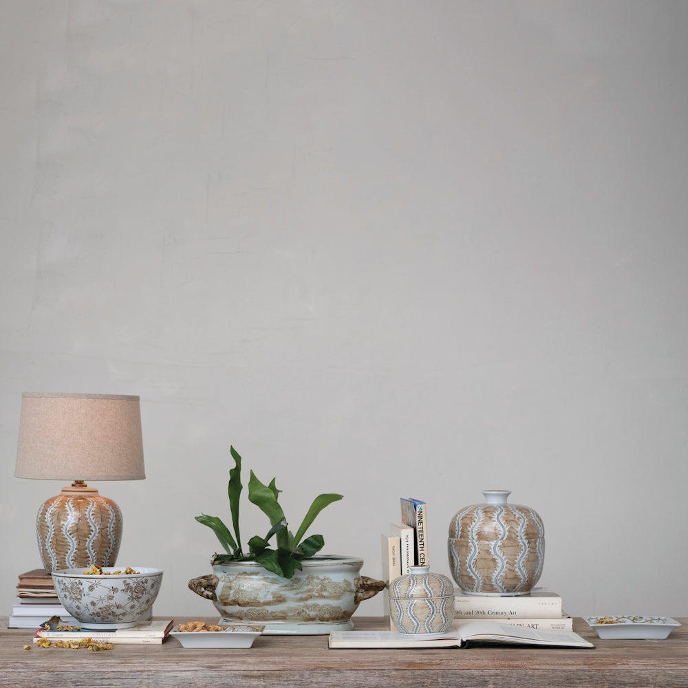 A tabletop display with books, a lamp, ceramic jars, and a green plant in a landscape-print planter.