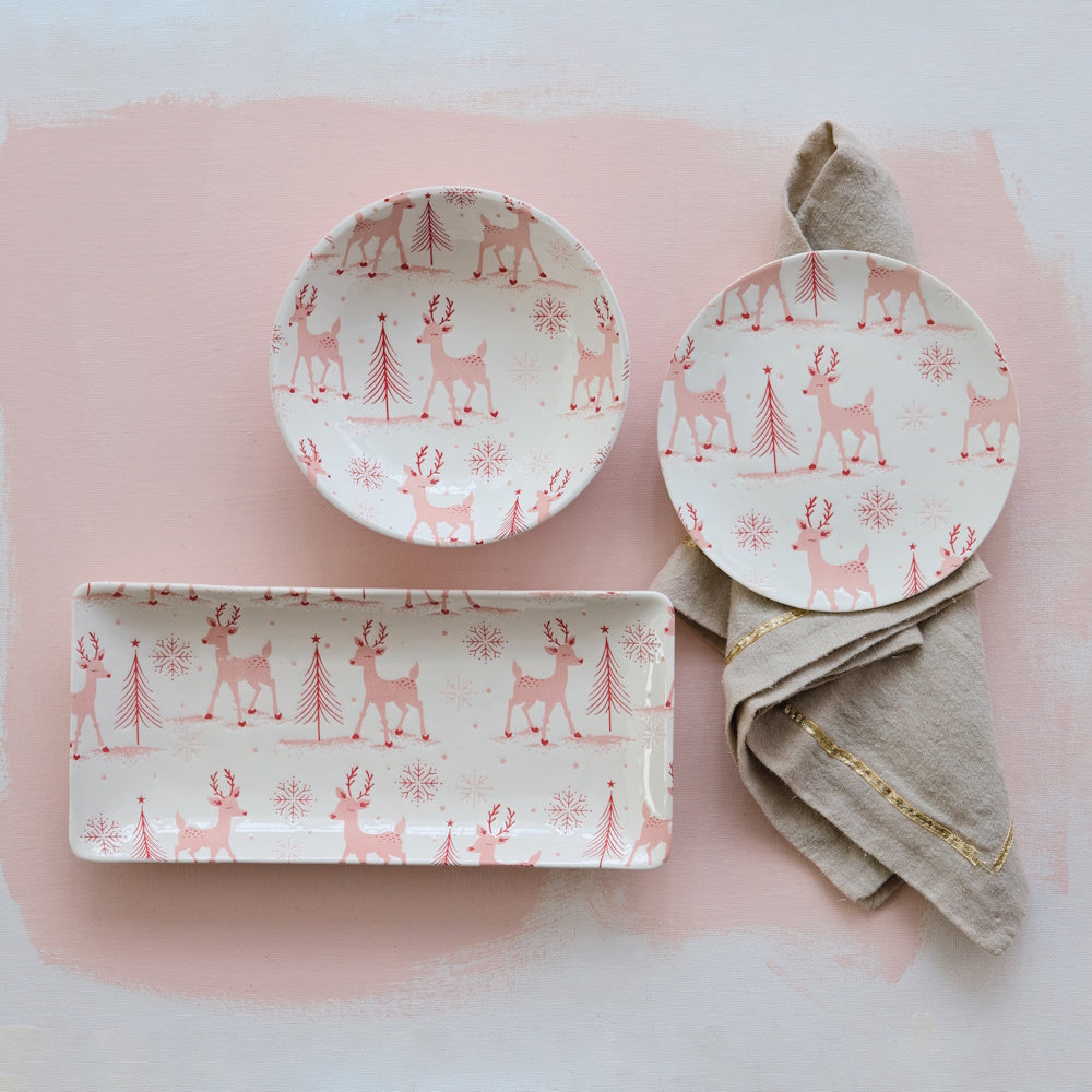 A bowl, plate, and platter featuring a holiday reindeer pattern, styled with a napkin on a pink background.