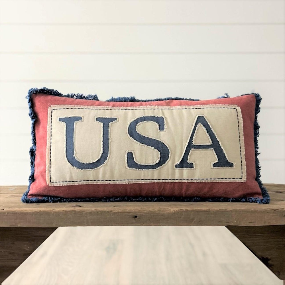 Rectangular red pillow with blue frayed edges and a center patch featuring the letters USA in blue.
