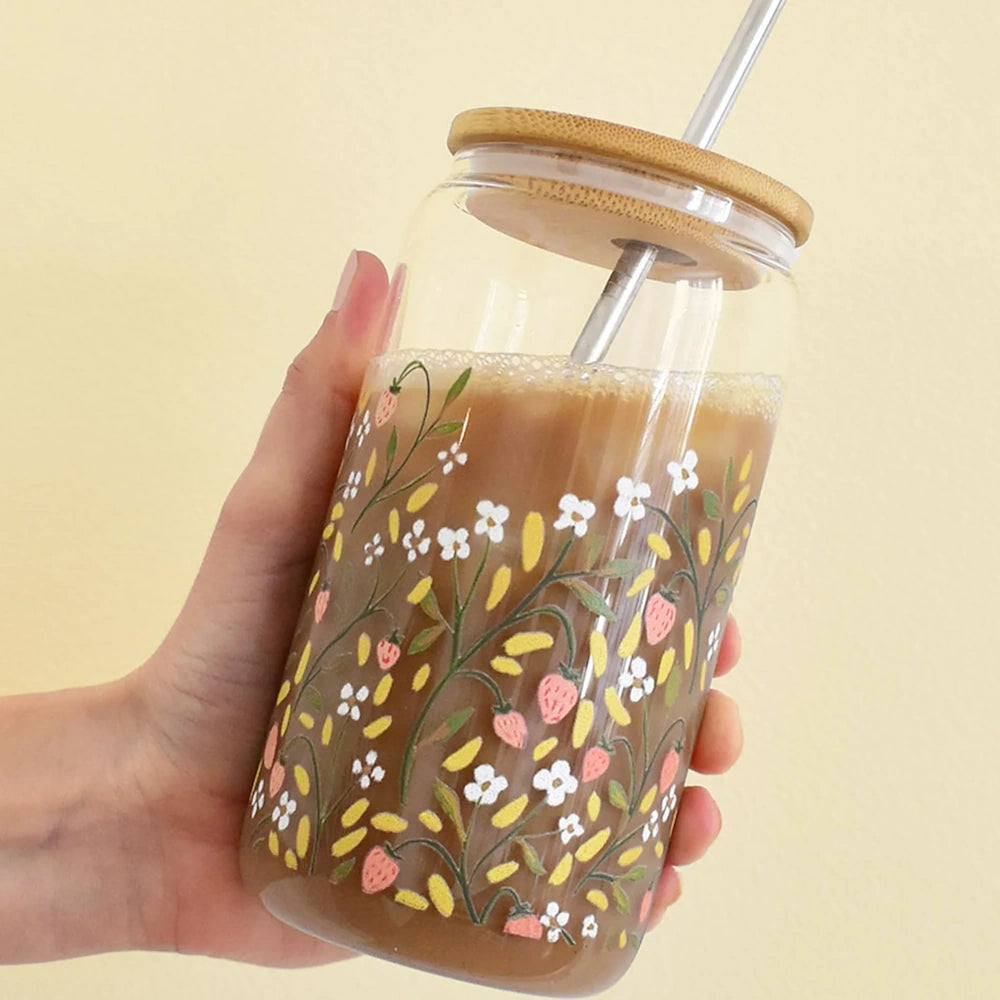 A hand holding a floral glass tumbler filled with iced coffee, featuring a bamboo lid and metal straw.