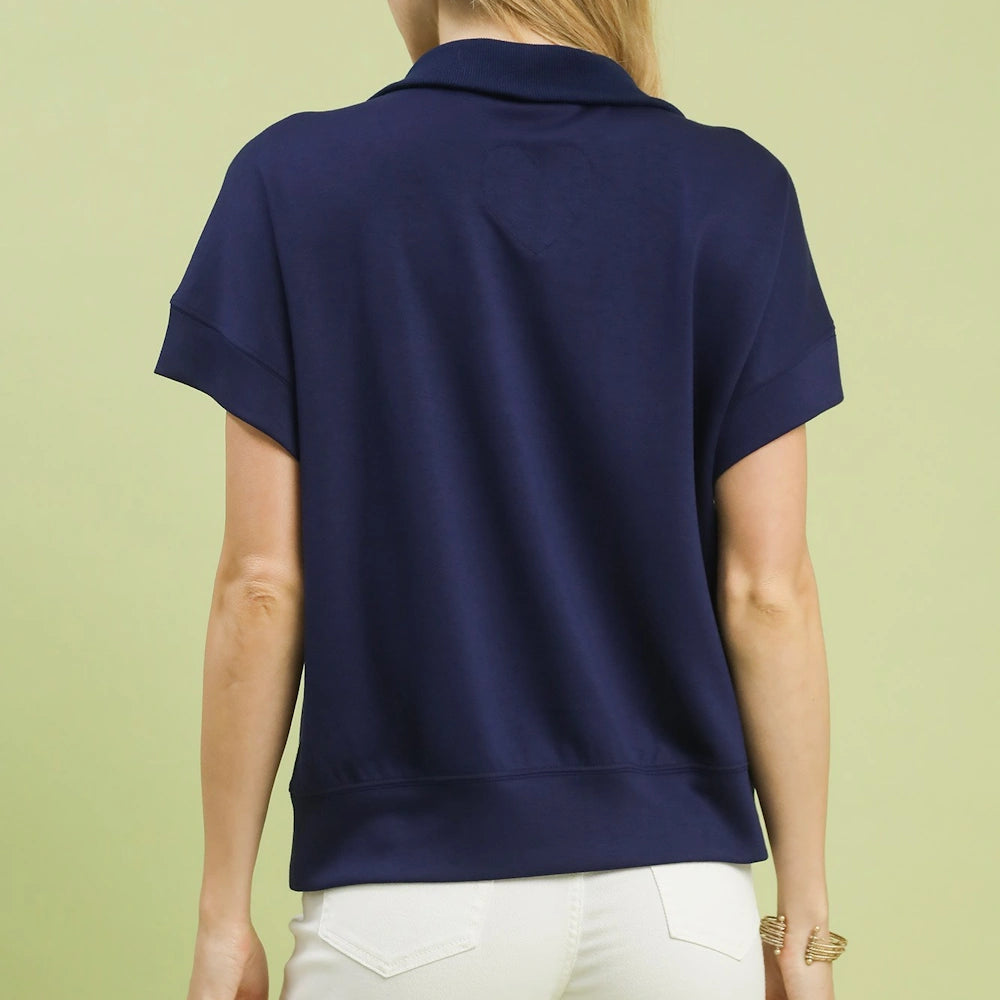 Back view of a navy blue short-sleeve knit polo shirt with a subtle heart-shaped stitch paired with white pants.