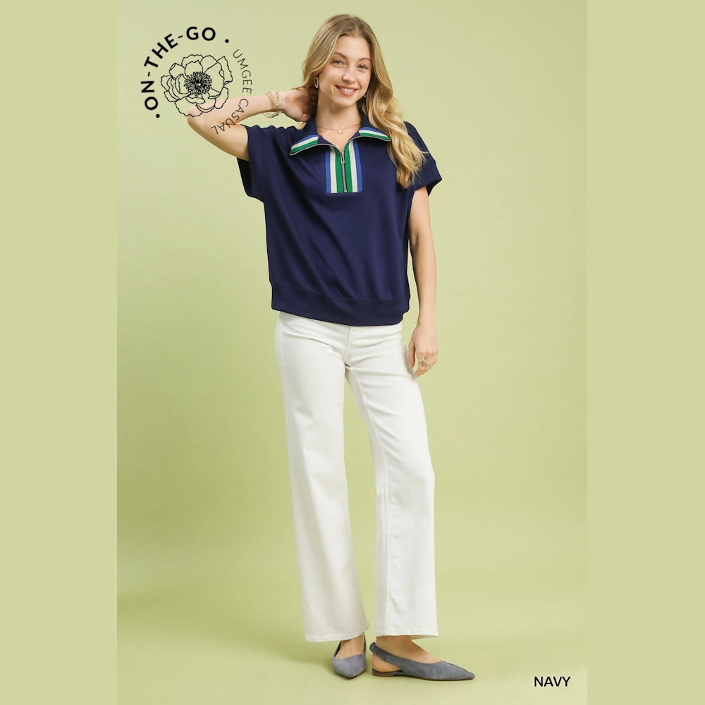 Woman in a navy half-zip knit top with green and white striped trim, paired with white pants and grey pointed flats.