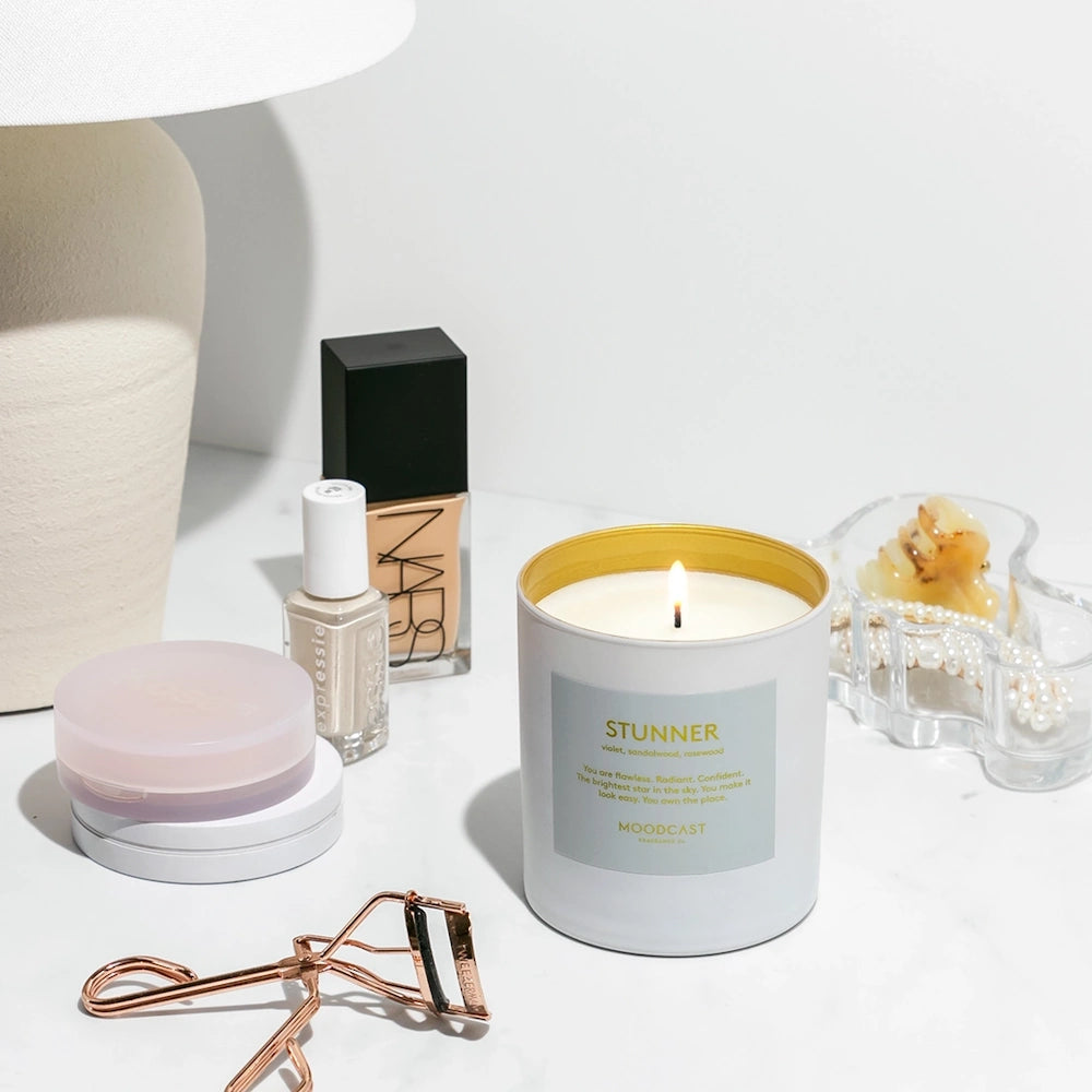 Lit white "Stunner" candle surrounded by makeup, nail polish, an eyelash curler, and accessories.