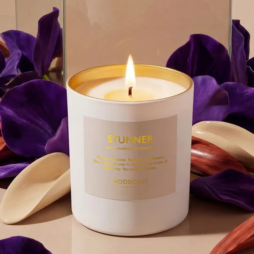 Lit Moodcast Stunner candle set against a backdrop of deep purple petals and smooth wooden accents.