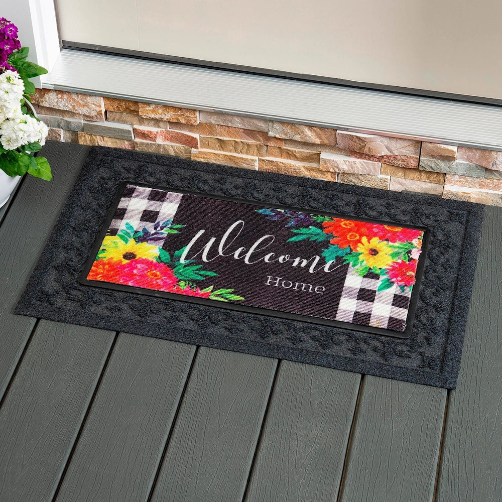 A welcome mat with colorful flowers and plaid patterns sits on a dark gray porch in front of a doorway.