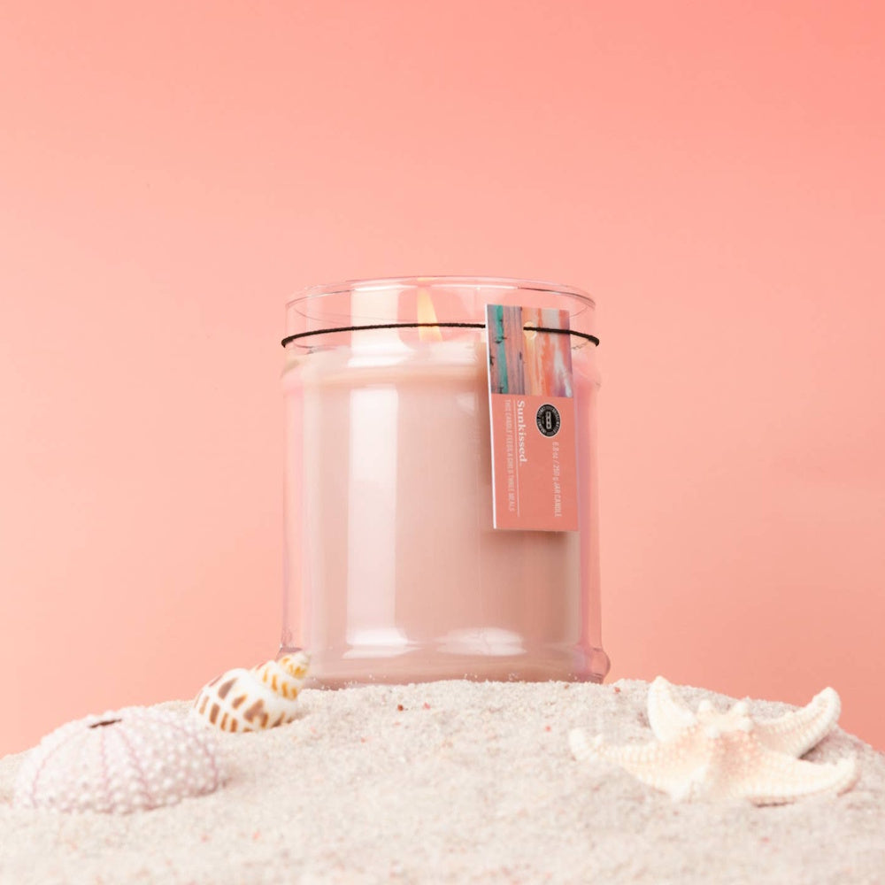 Bridgewater Candle Company Sunkissed jar candle on white sand with seashells and a starfish on a pink background.
