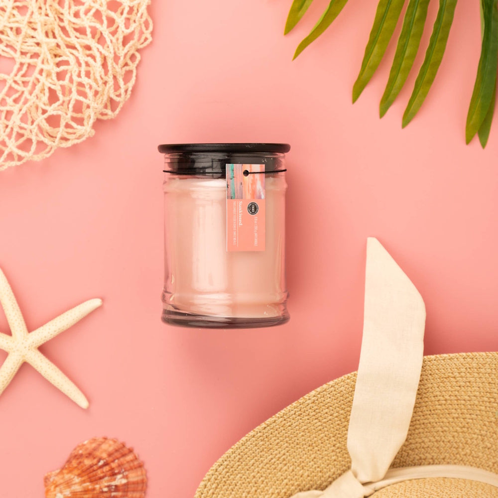 Sunkissed jar candle on pink background with starfish, seashell, palm leaf, and sun hat for summer home decor.