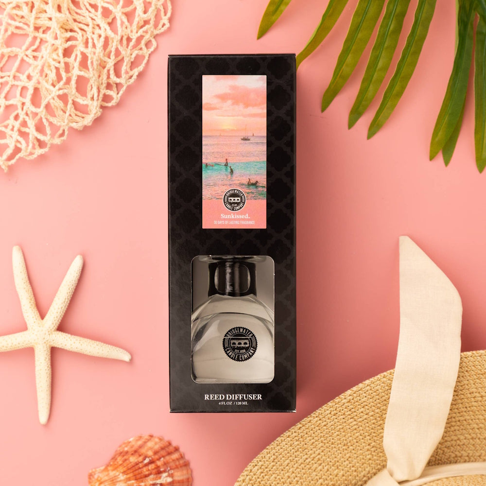 Sunkissed scented reed diffuser bottle and box styled with a starfish, seashells, and palm leaves on pink.