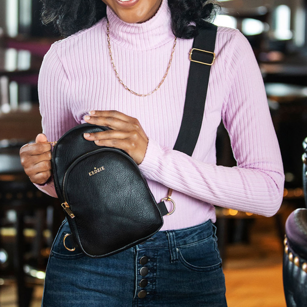 A person wearing a pink turtleneck and jeans, holding a black Kedzie sling bag across their chest.