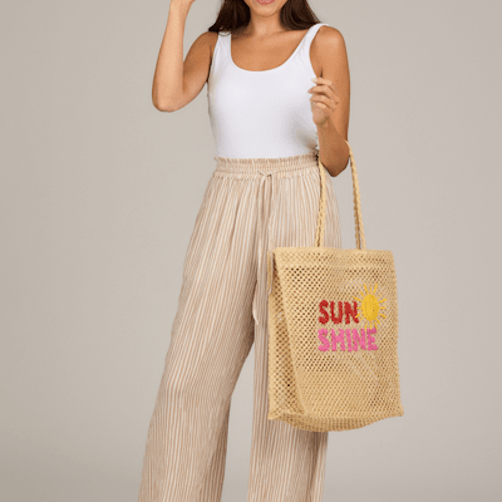 Woman holding a woven mesh tote bag that says "SUNSHINE" with a sun graphic, wearing striped wide-leg pants.