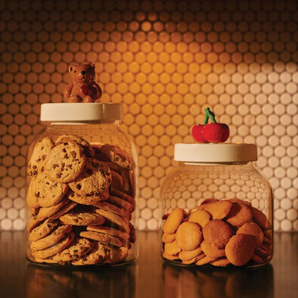 Two clear glass cookie jars with white lids, topped with a bear figurine and cherries, filled with cookies.