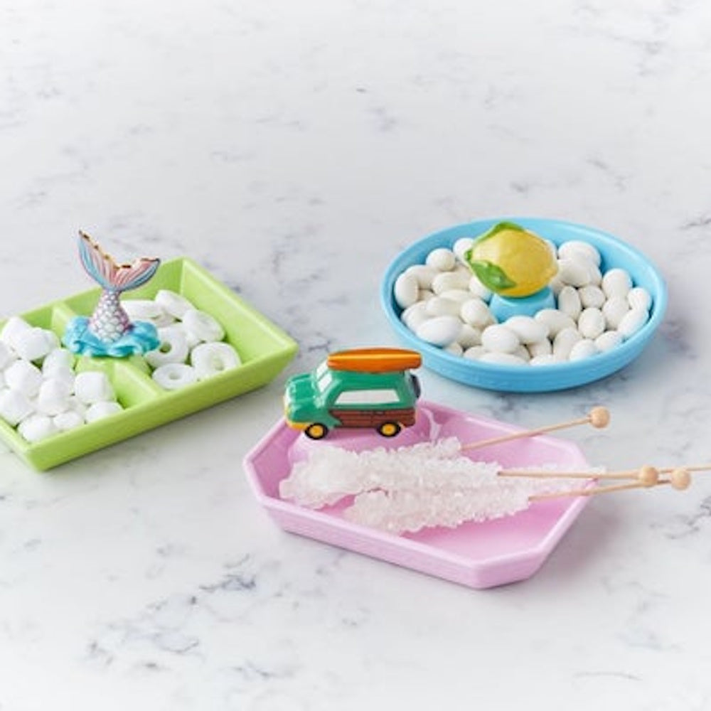 Three colorful beach-themed candy dishes filled with various sweets on a white marble surface.