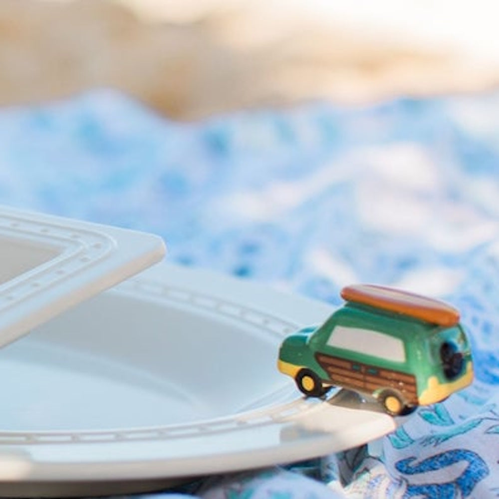 A toy surf van sits on the rim of a white plate over a blue patterned blanket.