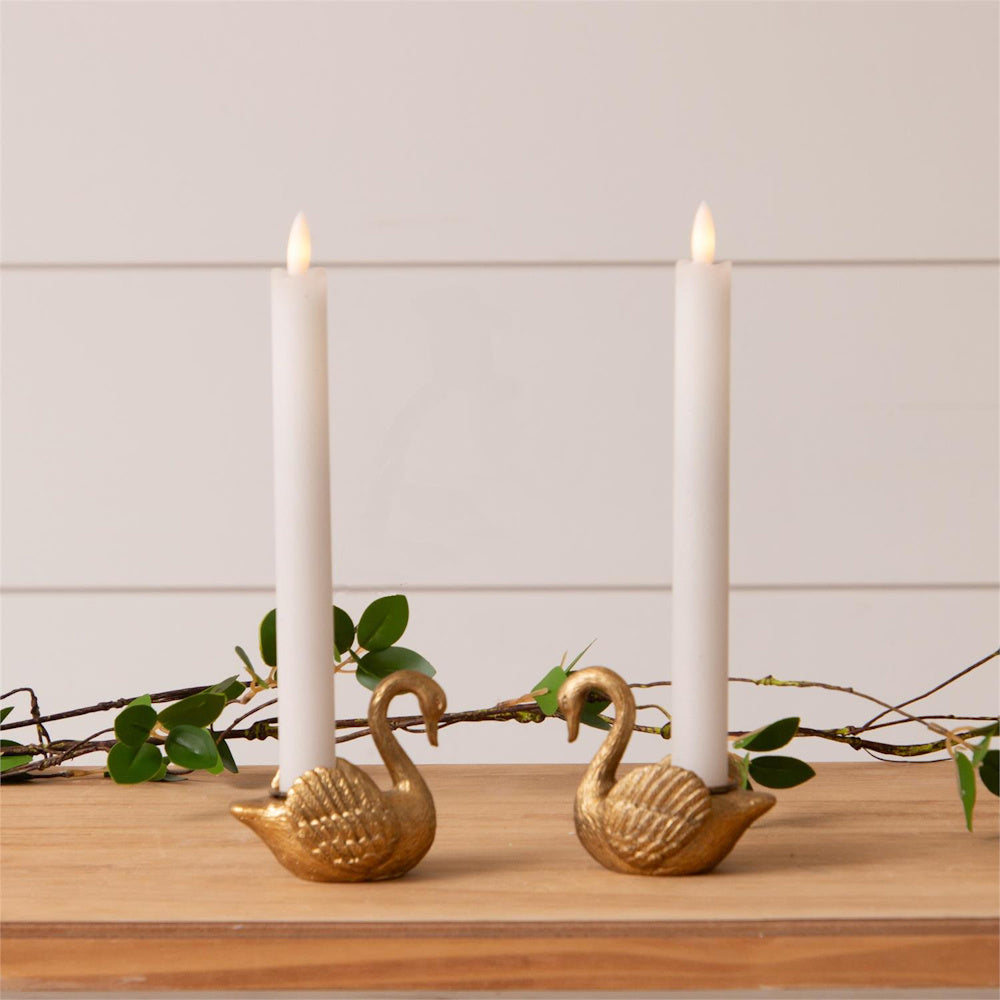 Gold swan tapered candle holders with white candles on a wooden surface with green leaves and a white paneled background.