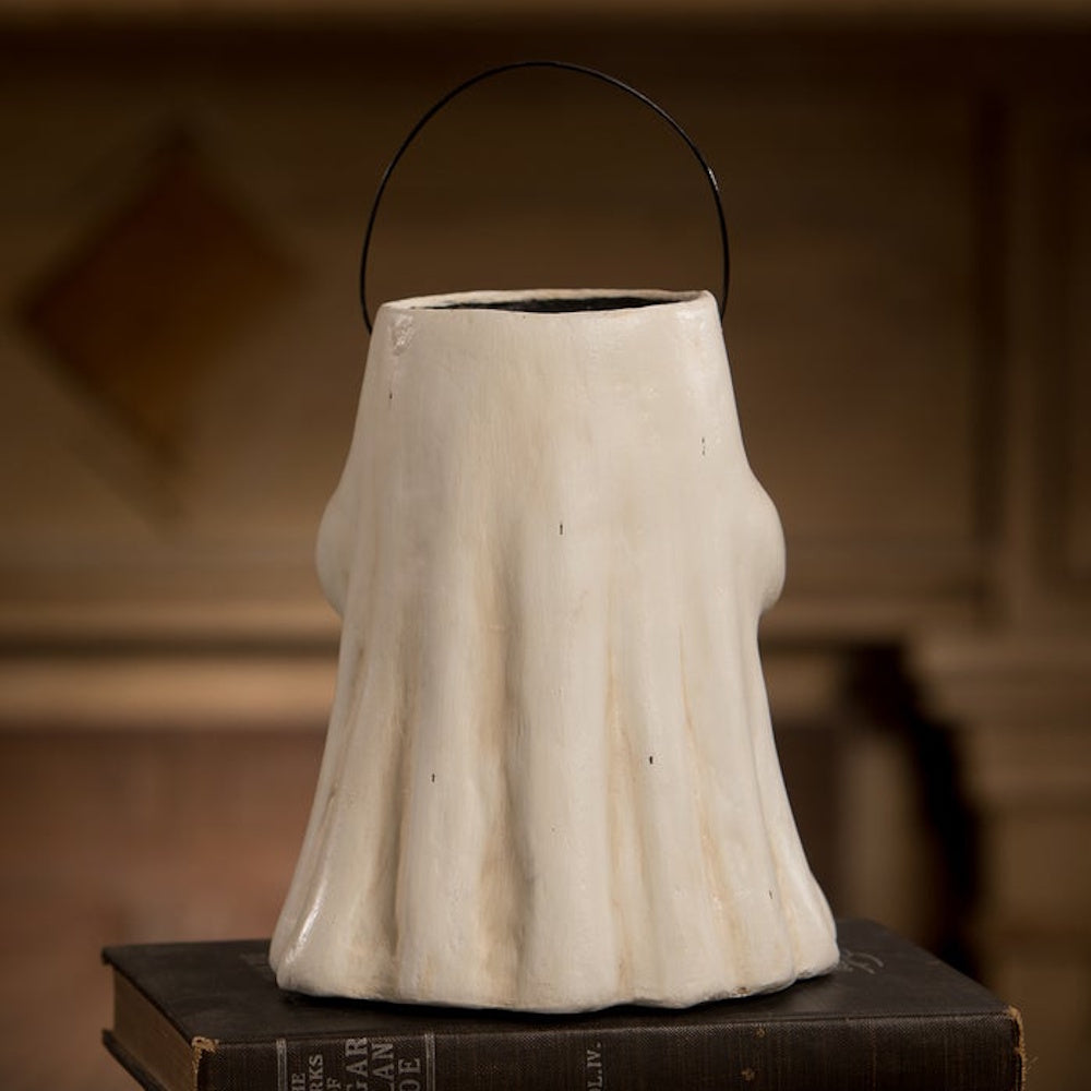 Back view of a white, paper-mache ghost figurine bucket with a black wire handle, sitting on a book.