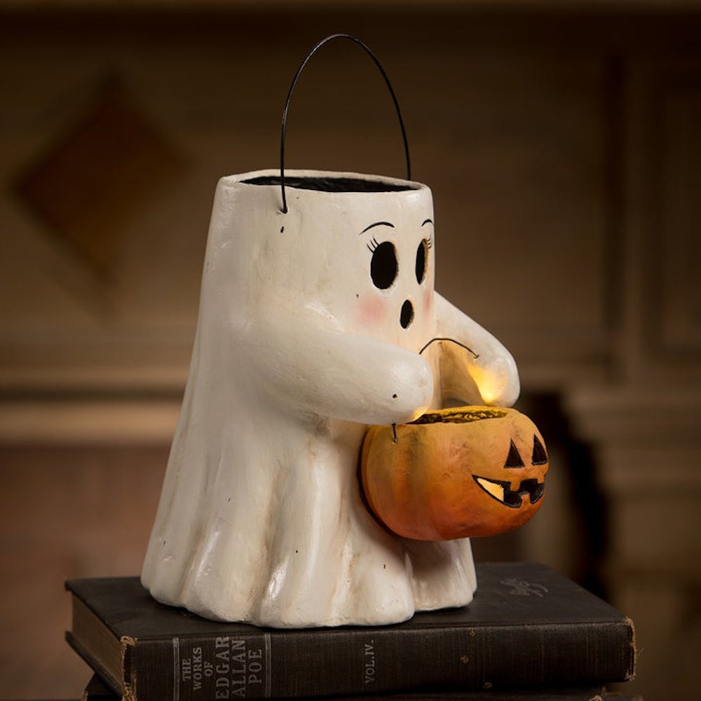 A paper-mache ghost figurine holding a glowing jack-o'-lantern bucket while sitting on an old book.