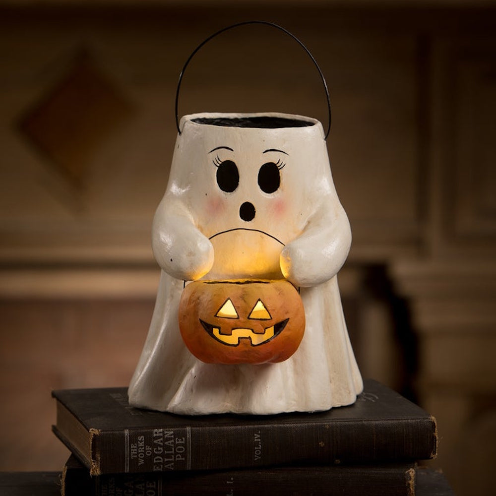 A paper mache ghost figurine sitting on old books, holding a glowing jack-o'-lantern pumpkin bucket.