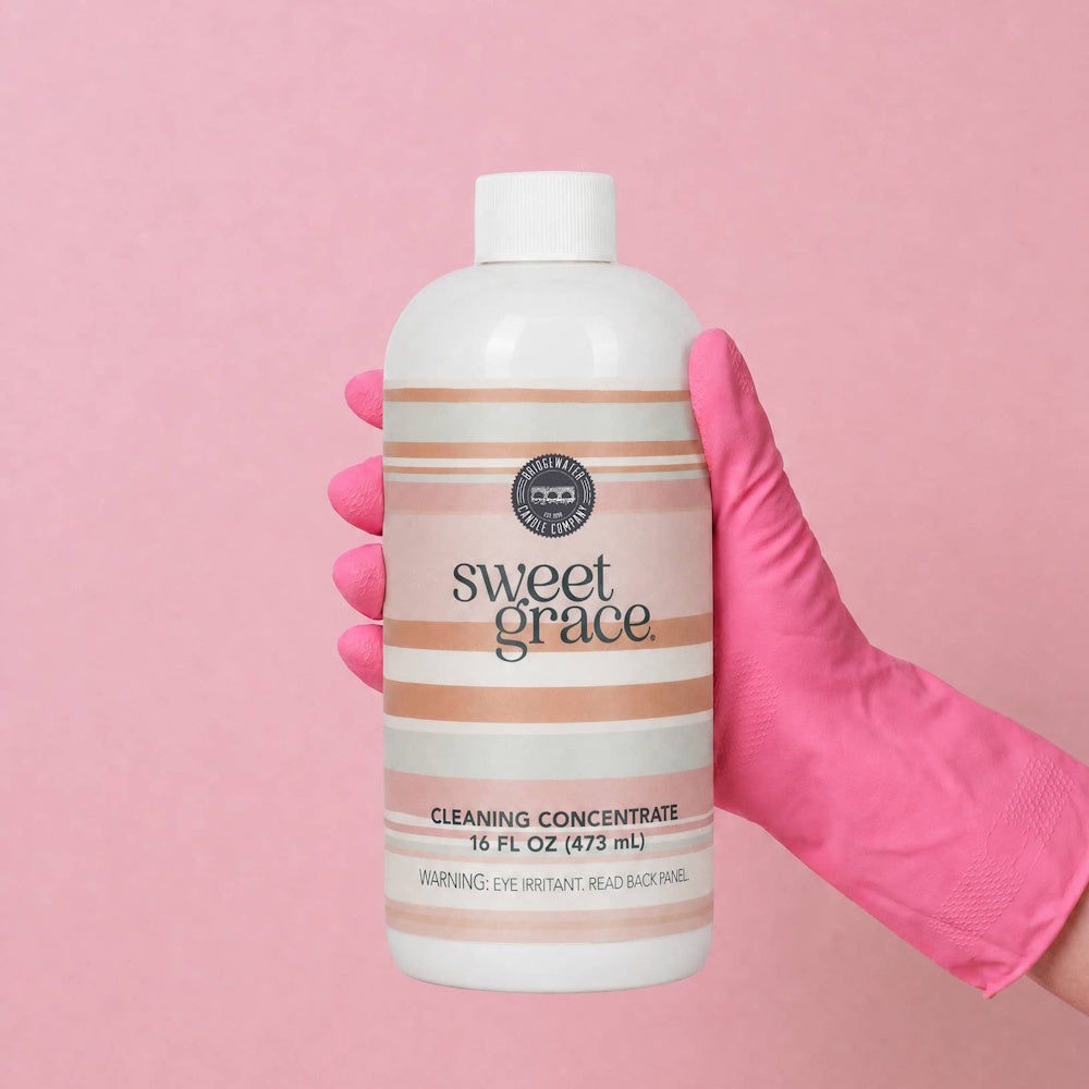 Gloved hand holding a bottle of Sweet Grace Cleaning Concentrate against a bright pink background.